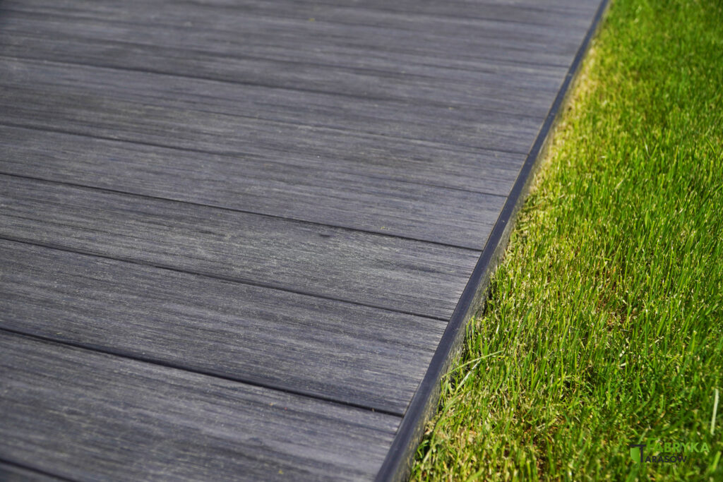 Szara deska kompozytowa ułożona w systemie tarasowym, w poprzek, z zastosowaniem listwy tarasowej. Nisko położony taras, po prawej stronie grafiki trawnik.