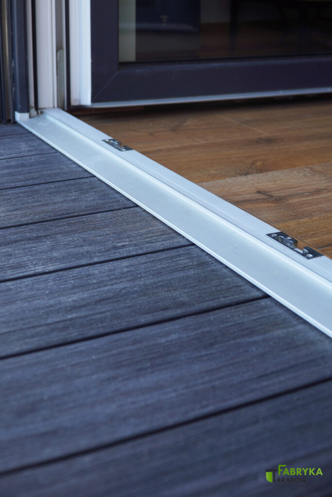 Zbliżenie na taras z desek kompozytowych w miejscu łączenia z parkietem domu przy wejściu. Deski kompozytowe są w kolorze ciemnym i równo łączą się z obramowaniem drzwi wejściowych.