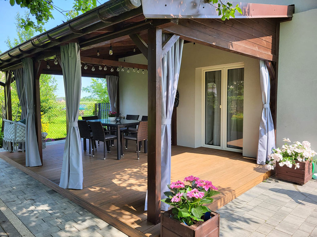 Aranżacja - zadaszony taras z zasłonami, umeblowaniem oraz roślinnością.