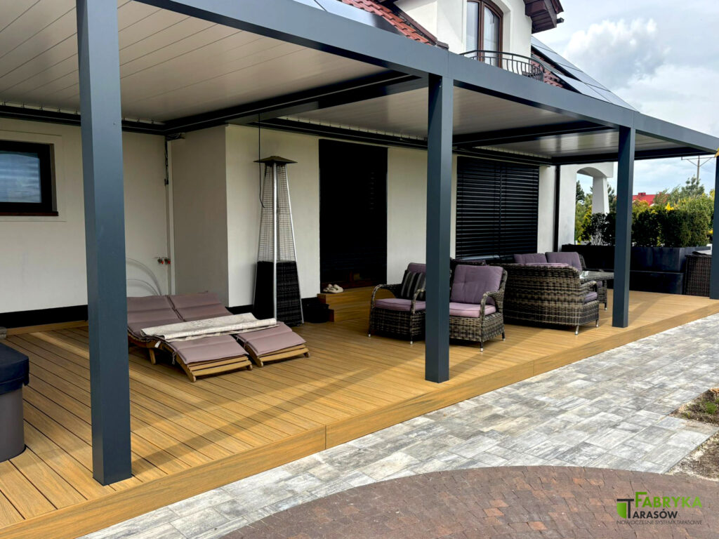 Szeroki, rozbudowany taras z deski kompozytowej, umieszczony przy domu wraz z zadaszeniem i umeblowaniem. Taras zrealizowany przez Fabrykę Tarasów.