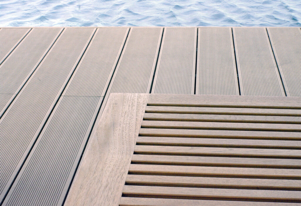 Drewniany stolik oraz przybrzeżny taras z deski kompozytowej. Stolik oraz deski kompozytowe są dobrane kolorystycznie. W tle, na górze obrazu widoczne fale wody.
