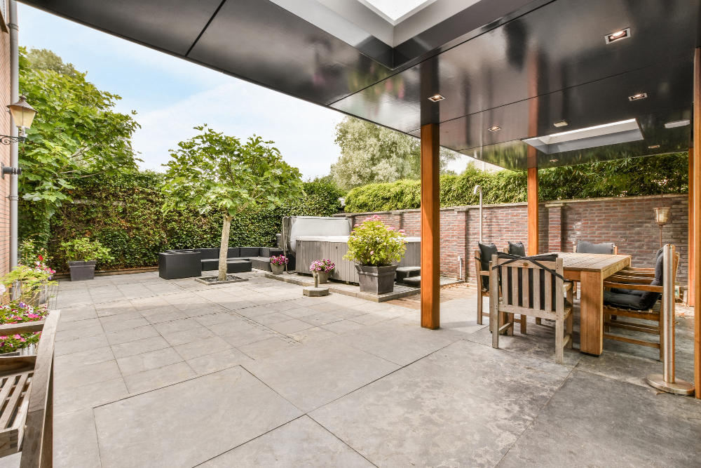 Taras z płyt betonowych oraz patio, wykonane z dwóch rodzajów płyt tarasowych. Taras jest zadaszony, widoczne w tle meble ogrodowe, jacuzzi oraz donice z roślinami.
